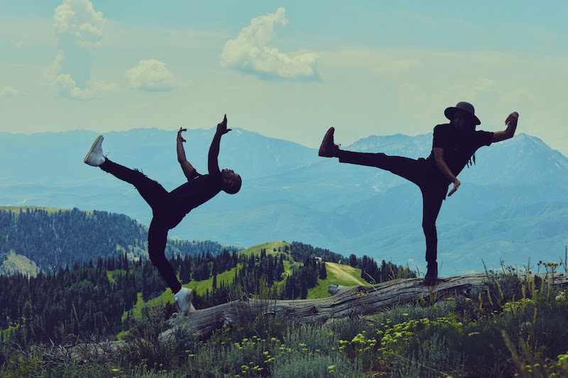 Two men in black casual clothes and sneakers kick their legs into their air on a hill. There's a scene of mountains and greenery behind them.